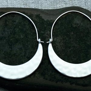 May include: A pair of silver-colored hoop earrings. Each earring features a crescent-shaped hammered metal design suspended from a thin, curved wire. The earrings are displayed on a dark, textured surface.