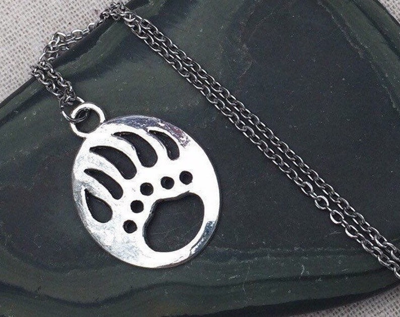 May include: A silver necklace with a bear paw print pendant. The pendant is a circle with a detailed outline of a bear paw print.