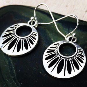 May include: A pair of silver-colored dangle earrings. Each earring features a circular design with a central hole and radiating, elongated cutouts. The earrings have a hook closure and a small ball detail near the top.