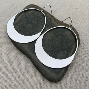 Puede incluir: Un par de pendientes de aro grandes, de color plateado. Cada pendiente tiene un dise&ntilde;o plano en forma de media luna en la mitad inferior. Los pendientes se muestran sobre una piedra gris oscura, sobre un fondo de tela neutra.