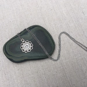 Puede incluir: Collar plateado con un colgante floral. El colgante tiene un dise&ntilde;o calado detallado con un elemento circular central y bordes festoneados. La fina cadena descansa sobre una piedra ovalada verde oscuro. Una joya delicada.