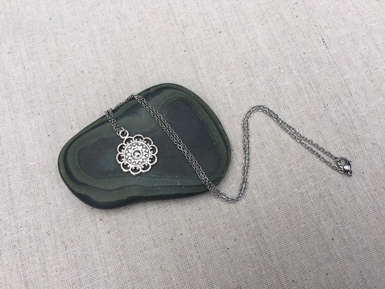 Puede incluir: Collar plateado con un colgante floral. El colgante tiene un dise&ntilde;o calado detallado con un elemento circular central y bordes festoneados. La fina cadena descansa sobre una piedra ovalada verde oscuro. Una joya delicada.