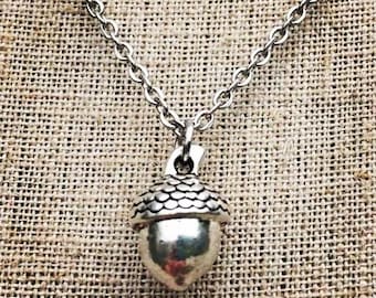 Puede incluir: Un collar con colgante de bellota plateada. El colgante de bellota tiene una tapa texturizada y un cuerpo liso y redondeado. La cadena del collar es de estilo eslabones delicados. El fondo es una tela texturizada de tono neutro.