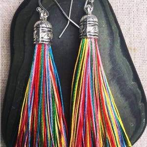 May include: A pair of multi-coloured tassel earrings. Each earring has a silver-toned cap with a decorative design and a cascade of vibrant threads in red, yellow, blue, and green. The earrings are displayed on a dark, textured surface.