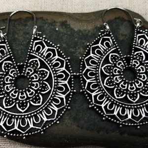 May include: A pair of silver hoop earrings with a mandala design. The earrings are large and have a delicate, intricate pattern.