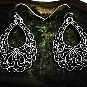 May include: A pair of silver-toned metal earrings featuring a teardrop-shaped filigree design with intricate swirling patterns and a central open cutout. Each earring is suspended from a curved metal hook wire for attachment to the earlobe.