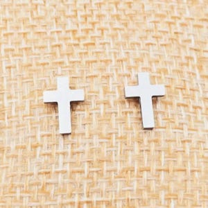 Op de afbeelding: Twee zilverkleurige kruisvormige oorstekers. De oorbellen worden gepresenteerd op een getextureerd, beige oppervlak. De kruizen hebben een eenvoudig, minimalistisch ontwerp, met strakke lijnen en een gepolijste afwerking.