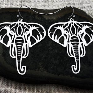 May include: A pair of white elephant-shaped earrings. The earrings are cut out with intricate details, including the elephant's head, ears, and trunk. The earrings are displayed on a dark, textured surface.