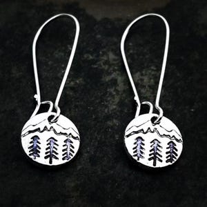 May include: A pair of silver-toned dangle earrings. Each earring features a circular pendant with a mountain range design above three stylized trees. The trees have a dark blue fill. The earrings have long, curved ear wires.