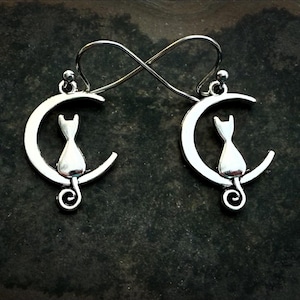 May include: Silver-toned cat earrings. Each earring features a crescent moon shape with a cat silhouette perched on it. The earrings are connected by a figure-eight shaped wire. The cat's tail curls into a spiral.