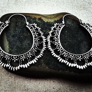 May include: A pair of silver-colored hoop earrings with an intricate floral and geometric openwork design. The earrings are displayed on a dark, textured stone against a neutral fabric background. The earrings have a unique, bohemian style.