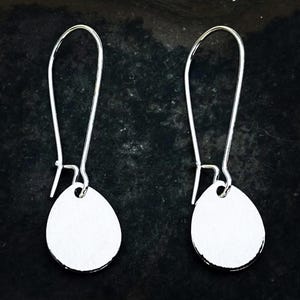May include: A pair of silver-toned drop earrings. Each earring features a long, curved wire hook and a small, oval-shaped pendant. The earrings are displayed against a dark, textured background.