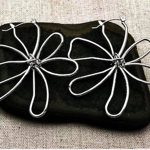 May include: A pair of silver-colored hoop earrings. Each earring features a flower design with six curved petals. The earrings are made of a shiny, metallic material and are displayed on a dark, textured surface.