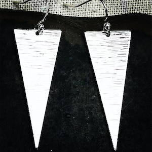 May include: A pair of silver-colored, triangular earrings with a textured surface. The earrings are suspended from silver-colored fish hook ear wires. The earrings are set against a dark background.