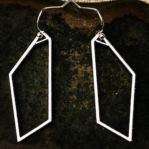 May include: A pair of silver-toned earrings featuring an open, geometric, asymmetrical pentagonal frame. Each earring is suspended from a simple silver-toned hook wire, showcasing a clean, hollow, angular metal design with a polished finish.