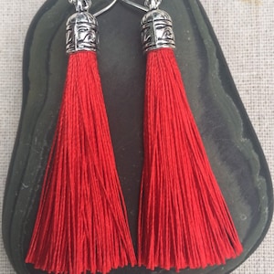 May include: A pair of red tassel earrings with silver metal tops. The tassels are long and flowing, and the metal tops have a decorative design.