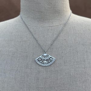 May include: Silver-toned pendant necklace featuring a fan-shaped charm. The charm has an intricate design with floral and geometric patterns. The necklace is displayed on a mannequin, showcasing the pendant's details and the chain's length.