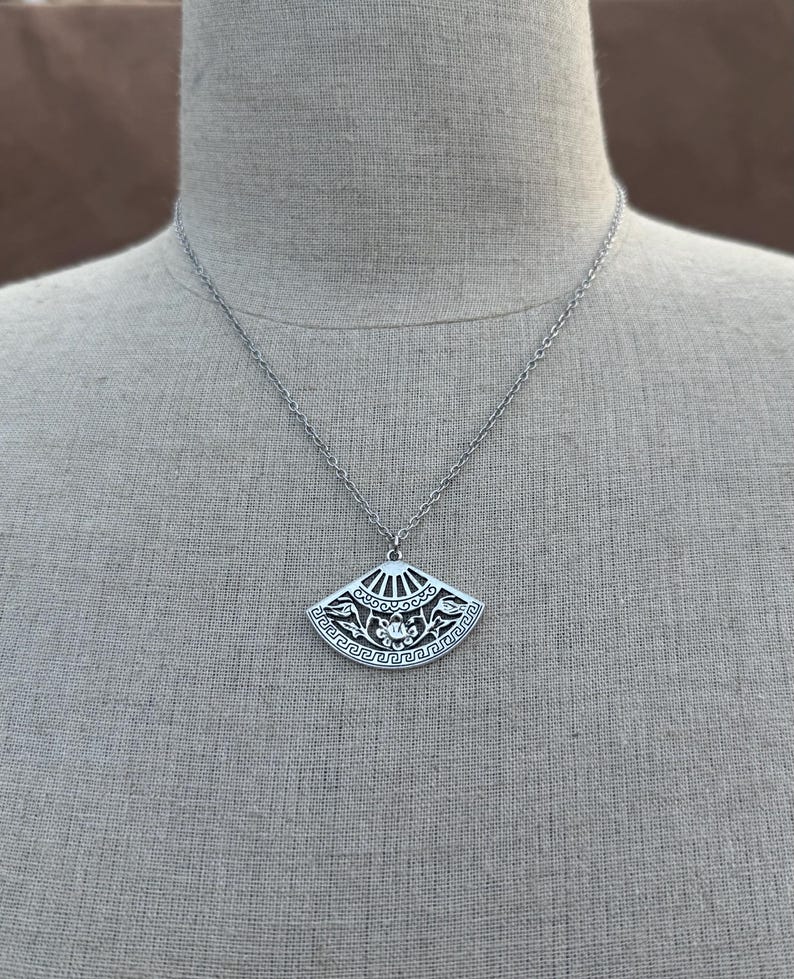 May include: Silver-toned pendant necklace featuring a fan-shaped charm. The charm has an intricate design with floral and geometric patterns. The necklace is displayed on a mannequin, showcasing the pendant's details and the chain's length.