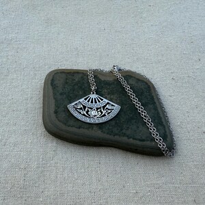 May include: A silver-toned pendant necklace featuring a fan-shaped design with intricate floral and geometric patterns. The pendant is suspended from a delicate chain. The necklace rests on a dark green stone against a neutral linen background.