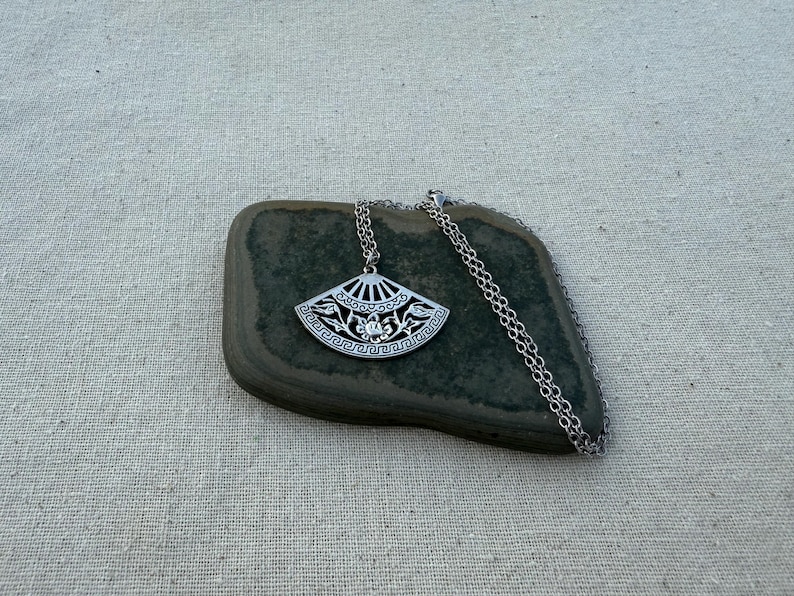 May include: A silver-toned pendant necklace featuring a fan-shaped design with intricate floral and geometric patterns. The pendant is suspended from a delicate chain. The necklace rests on a dark green stone against a neutral linen background.