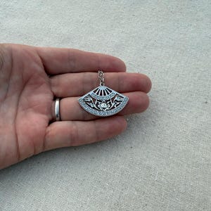 May include: A silver-colored pendant necklace featuring a fan-shaped design. The pendant has intricate cut-out patterns and a small chain. The fan design includes floral and geometric elements. The necklace is held in a hand.