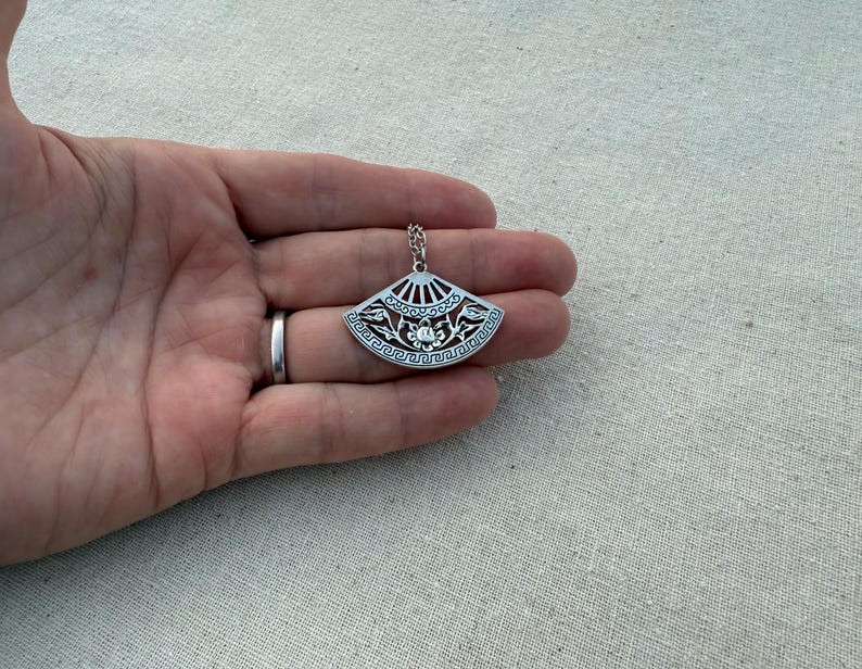May include: A silver-colored pendant necklace featuring a fan-shaped design. The pendant has intricate cut-out patterns and a small chain. The fan design includes floral and geometric elements. The necklace is held in a hand.
