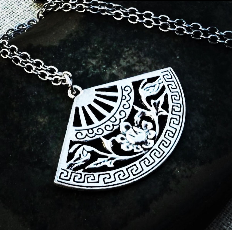 May include: Silver-toned pendant necklace featuring a fan-shaped design. The pendant showcases a detailed floral and geometric pattern with a sunburst design. The necklace has a delicate chain.