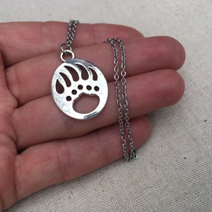 May include: A silver necklace with a circular pendant featuring a bear paw print design. The pendant is hollow and has a detailed outline of the paw print.