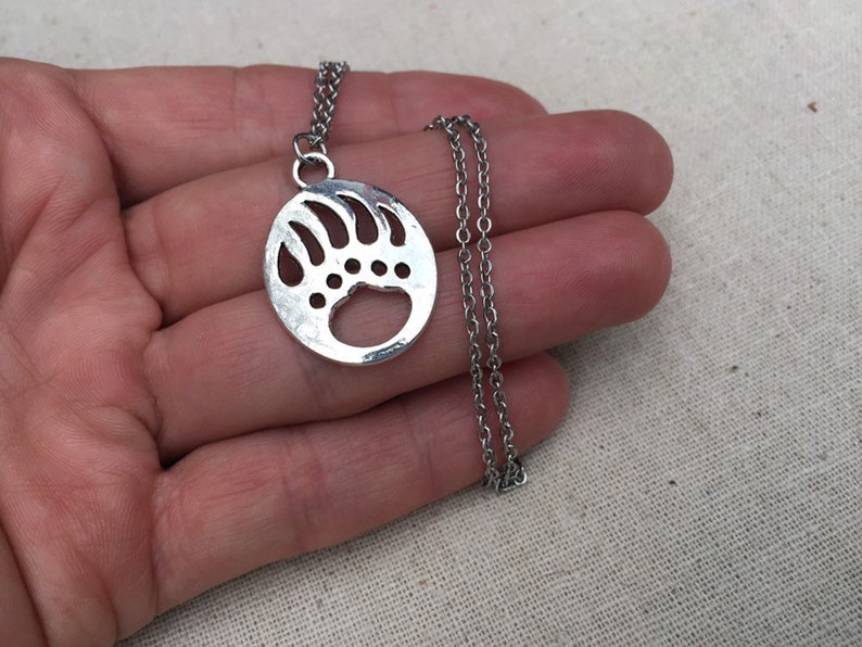 May include: A silver necklace with a circular pendant featuring a bear paw print design. The pendant is hollow and has a detailed outline of the paw print.