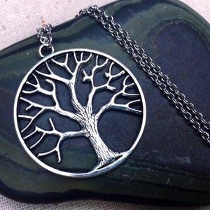 May include: A silver-toned metal pendant necklace featuring a circular frame containing a detailed tree silhouette with spreading branches and a textured trunk. The pendant is attached to a matching silver-toned link chain that rests in a loop.