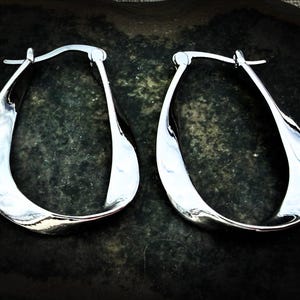 May include: A pair of polished silver hoop earrings. The earrings have a twisted, teardrop shape and a shiny, reflective surface. The earrings are displayed on a dark, textured surface.