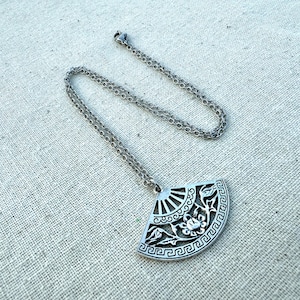 May include: Silver-toned pendant necklace featuring a fan-shaped pendant with intricate floral and geometric designs. The pendant hangs from a delicate chain, creating an elegant accessory. The necklace is displayed on a neutral linen surface.