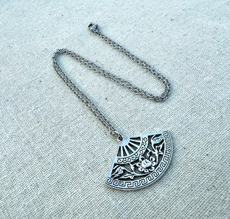 May include: Silver-toned pendant necklace featuring a fan-shaped pendant with intricate floral and geometric designs. The pendant hangs from a delicate chain, creating an elegant accessory. The necklace is displayed on a neutral linen surface.