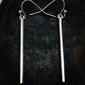May include: A pair of silver-colored, minimalist earrings. Each earring features a long, slender bar suspended from a curved ear wire with a small ball accent. The earrings are displayed against a dark, textured background.