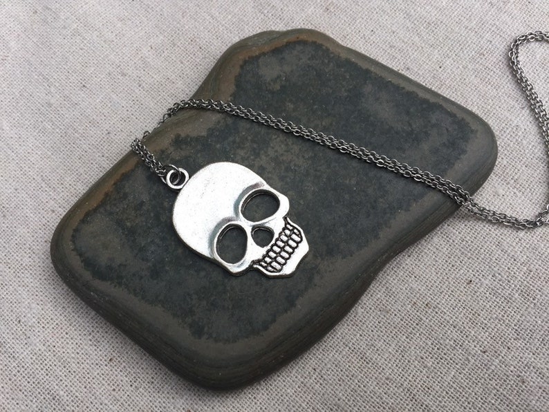 May include: A silver skull pendant necklace on a silver chain. The skull is detailed with eye sockets, a nose, and a mouth.
