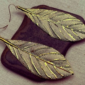 May include: A pair of gold-coloured leaf earrings. The earrings feature detailed textured veins and a hook closure. They are displayed on a dark, textured surface against a neutral background.