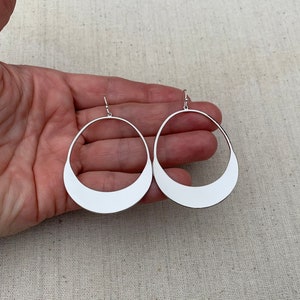 Puede incluir: Un par de pendientes de aro abiertos de color plateado. Cada pendiente tiene forma de l&aacute;grima con una mitad inferior plana y reflectante y una parte superior fina y curvada. Los pendientes se muestran sobre un fondo neutro.
