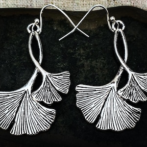 May include: A pair of silver-colored earrings featuring a ginkgo leaf design. The earrings have a hook closure and a detailed, fan-shaped leaf pattern. The earrings are displayed on a dark, textured surface.