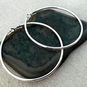 May include: A pair of polished silver-tone hoop earrings featuring a thin, circular wire design with a hinged clasp closure. The earrings are circular in shape with a smooth, reflective metallic finish and a simple, clean aesthetic for everyday wear.