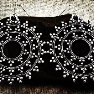 May include: A pair of silver-toned spiral earrings. Each earring features a circular design with a spiral pattern and small, round accents. The earrings are displayed on a dark surface against a textured background.