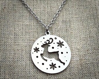 Op de afbeelding: Een zilverkleurige ketting met een cirkelvormige hanger. De hanger heeft een uitgesneden ontwerp van een rendier en sneeuwvlokken. Het rendier staat in profiel en de hanger hangt aan een ketting.