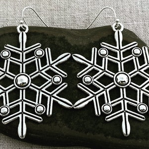 May include: A pair of silver snowflake earrings with a detailed design and small round accents. The earrings are hanging from a silver hook.