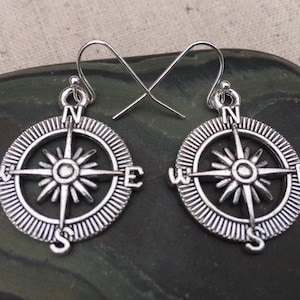 May include: A pair of silver compass earrings with a detailed design. The compass features the cardinal directions: North, South, East, and West.