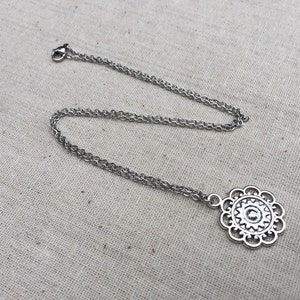 Puede incluir: Un collar plateado con un colgante floral. El colgante tiene un dise&ntilde;o calado detallado con un elemento circular central y formas de p&eacute;talos. La cadena es de estilo fino y enlazado.