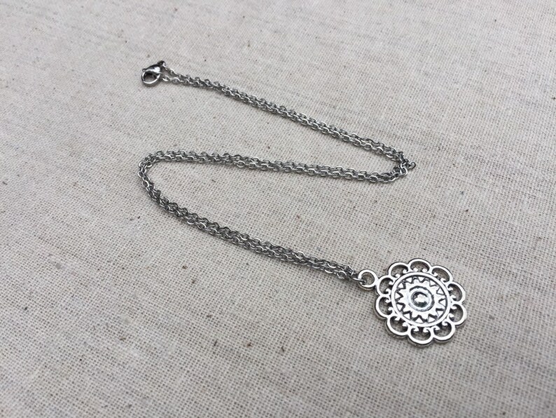 Puede incluir: Un collar plateado con un colgante floral. El colgante tiene un dise&ntilde;o calado detallado con un elemento circular central y formas de p&eacute;talos. La cadena es de estilo fino y enlazado.