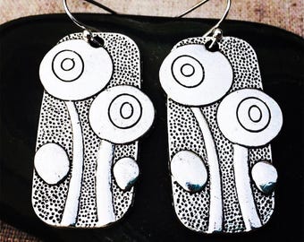 May include: A pair of silver-toned earrings featuring a floral design. Each earring has a rectangular shape with a dotted background and three circular flower-like shapes. The earrings have a hook closure.