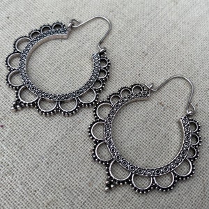 May include: A pair of silver-toned hoop earrings. Each earring features a circular design with a scalloped edge and intricate detailing. The earrings have a hook closure and a decorative, dangling element at the bottom.