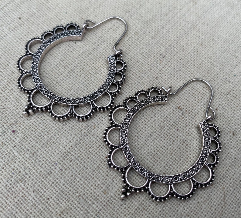 May include: A pair of silver-toned hoop earrings. Each earring features a circular design with a scalloped edge and intricate detailing. The earrings have a hook closure and a decorative, dangling element at the bottom.