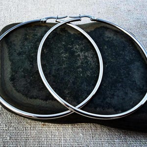 May include: A pair of large, silver hoop earrings. The earrings are circular and have a polished, reflective surface. They are displayed on a dark, textured stone against a neutral fabric background. The earrings are a classic jewelry accessory.