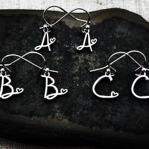 May include: Silver initial earrings with the letters A, B, and C, each accompanied by a small heart. The earrings are presented on a dark, textured surface.